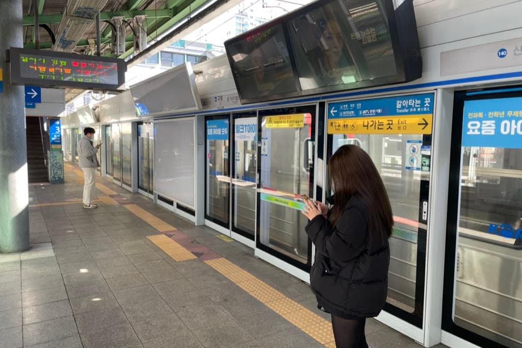 Quai de métro aérien aux portes palières vitrées, une voyageuse consulte son téléphone