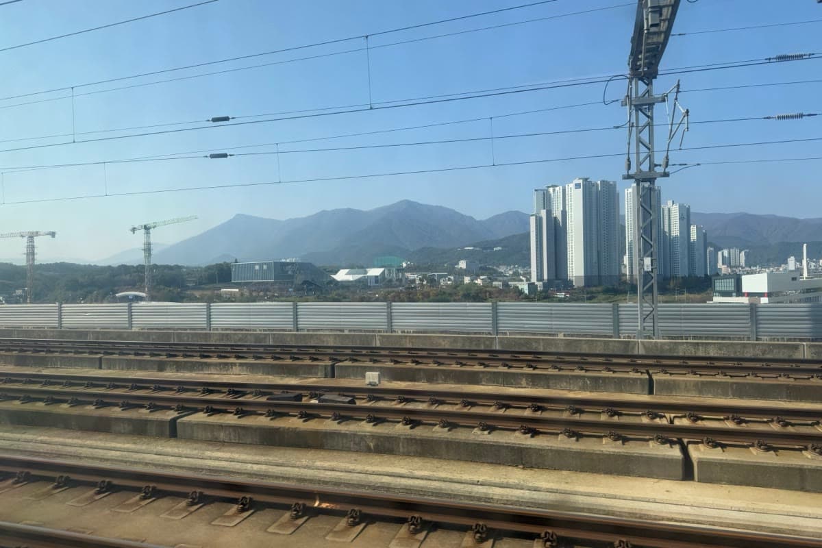 Voies ferrées du KTX au pied d'immeubles et de montagnes, sous un ciel bleu